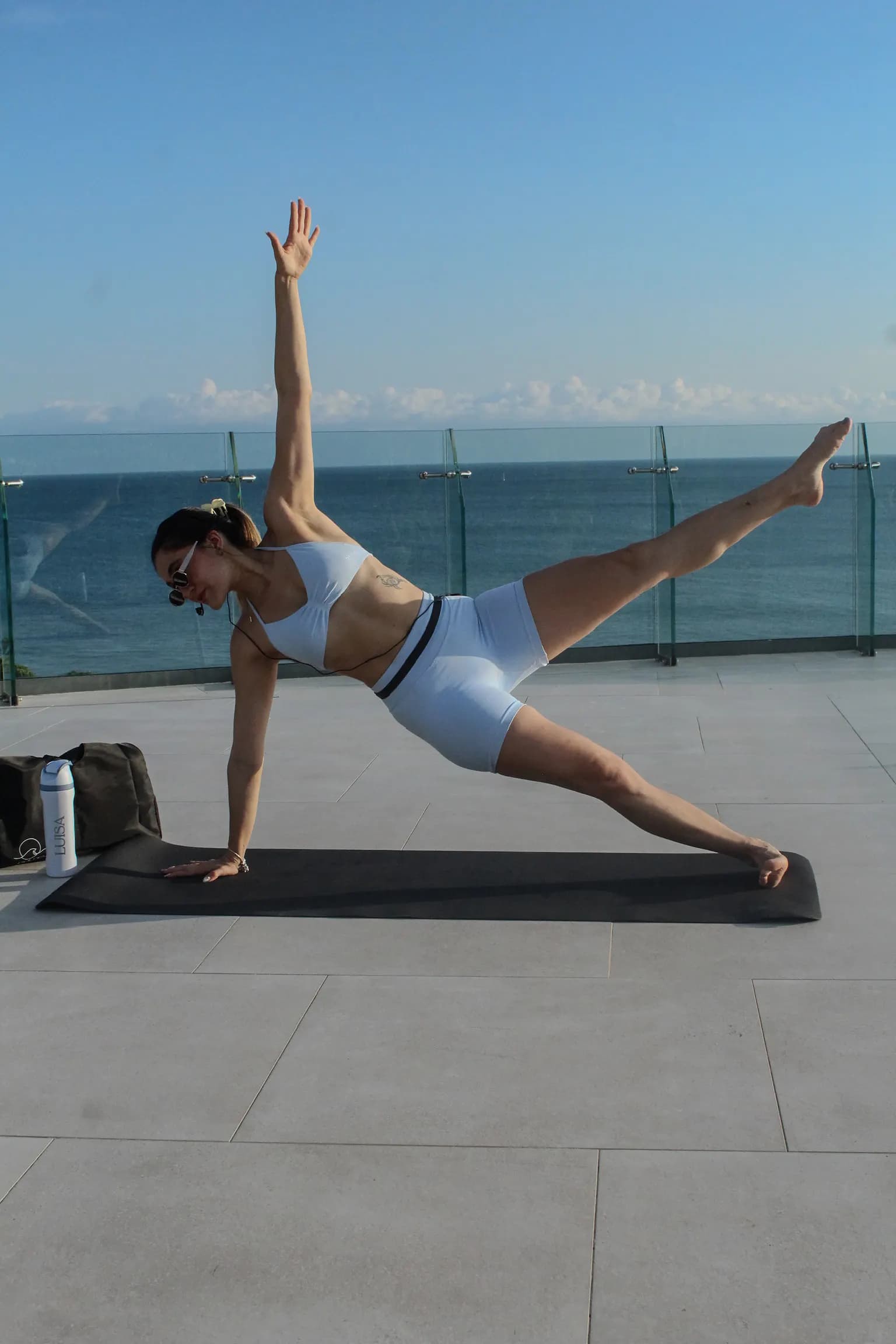 Luisa en plancha lateral con un brazo y una pierna en alto, en una terraza frente al mar